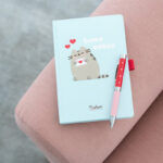 Notatnik Pusheen „Love Notes” z czerwonym długopisem, leżący na różowym pufie.