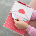 Notatnik Pusheen z czerwonym długopisem i kopertą z sercem trzymany w dłoniach.