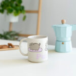 Kubek Pusheen z serii Purrito ustawiony obok błękitnego kawiarka na białym stole